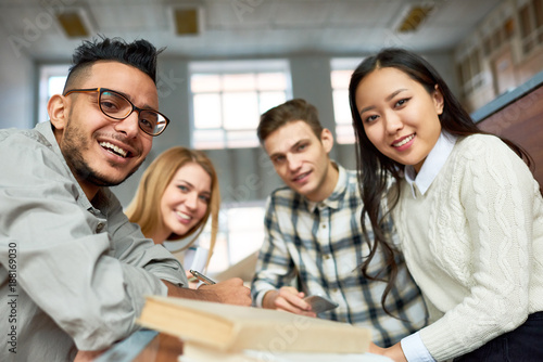 Фото: Multi-ethnic group of cheerful students looking at camera and smiling sitting at desk in lecture hall of modern college Multi-ethnic group of cheerful students looking at camera and smiling sitting at desk in lecture hall of modern college