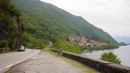 Cars driving along a mountain road along the shore of Lake Como, Italy with mountains and quaint village in the background.