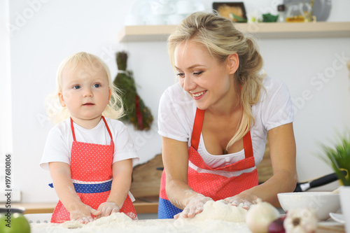 Plakat Mała dziewczynka i jej blondynki mama w czerwonych fartuchach bawić się i śmia się podczas gdy ugniatający ciasto w kuchni. Domowe ciasta na chleb, pizzę lub upiecz ciasteczka