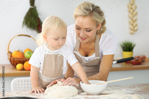 Plakat Mała dziewczynka i jej blondynki mama w czerwonych fartuchach bawić się i śmia się podczas gdy ugniatający ciasto w kuchni. Domowe ciasta na chleb, pizzę lub upiecz ciasteczka