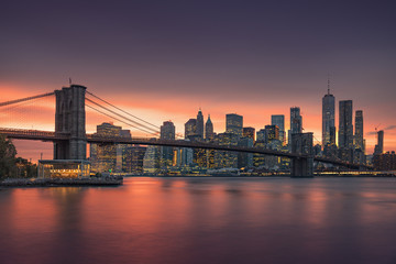 Famous Brooklyn Bridge in New York City with financial district - downtown Manhattan in background. Sightseeing boat on the East River and beautiful sunset over Jane's Carousel.