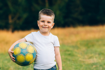 Child with a soccer ball under his arm at house garden with grass background, smiling. Kid Sport and Soccer World Cup Concept.