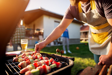 Dedicated man turning vegetables and meat on a stick to grill on other side as well while rest of the family is scattered around the house yard.
