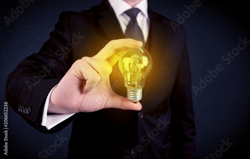 Фото: A business man in suit holding a glowing yellow light bulb in his hand while working in the office concept. A business man in suit holding a glowing yellow light bulb in his hand while working in the office concept.