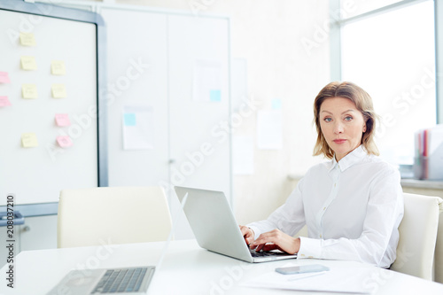 Фото: Portrait of confident mature businesswoman looking at camera while typing on laptop at office desk Portrait of confident mature businesswoman looking at camera while typing on laptop at office desk