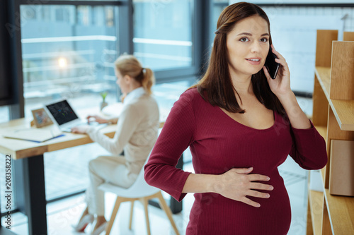 Фото: Expecting baby. Pretty female person standing on the foreground and putting right hand on the belly while calling to her doctor Expecting baby. Pretty female person standing on the foreground and putting right hand on the belly while calling to her doctor