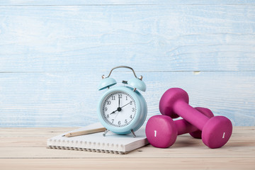Fitness concept with pink dumbbells, alarm clock and notepad for workout plan