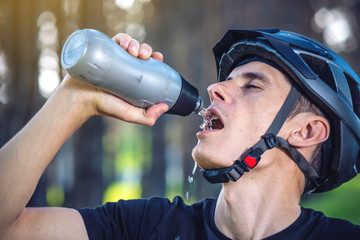 Cyclist in a helmet drinking water from a bottle while riding in the Park among the trees. Active and healthy lifestyle