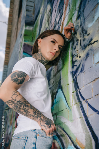 Фото: low angle view of stylish tattooed girl posing near wall with colorful graffiti low angle view of stylish tattooed girl posing near wall with colorful graffiti