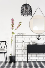 Lamp and mirror above black washbasin in modern bathroom interior with plant and chair. Real photo