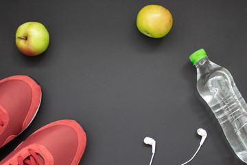 Fitness flat lay. Healthy lifestyle and sport concept. Red sneakers, white earphones, apples and bottle of water on a black background.