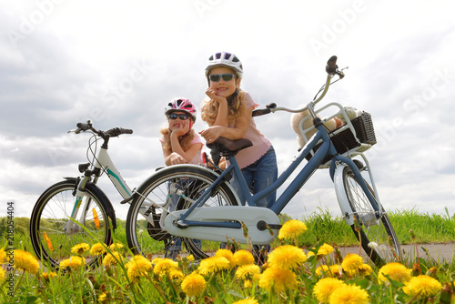Foto obraz na płótnie canvas jednoczęściowy - Twin sisters relax from riding their bicycles in front of a dandelion field. They have happy smiling faces on them as they enjoy the outdoor cycle tour.