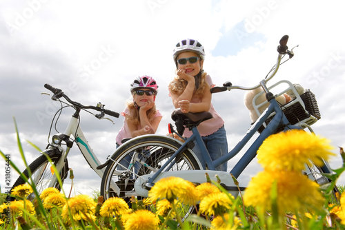 Foto obraz na płótnie canvas jednoczęściowy - Twin sisters relax from riding their bicycles in front of a dandelion field. They have happy smiling faces on them as they enjoy the outdoor cycle tour.