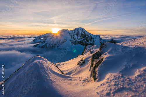 Foto obraz na płótnie canvas czteroczęściowy - "tetraptyk" - Stunning sunset or sunrise in winter alpine like snow landscape. Inversion, sun star peaking behind high rocky and icy summit. Purple, pink, blue and orange colors. Ladovy stit in winter High Tatras.