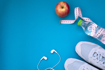 Fitness flat lay. Healthy lifestyle and sport concept. Blue sneakers, apple, bottle of water, tape measure and white earphones on a blue background.