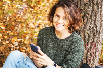 Beautiful young smiling  student girl woman with wavy hair sitting on grass and on fallen autumn leaves with smartphones in hands