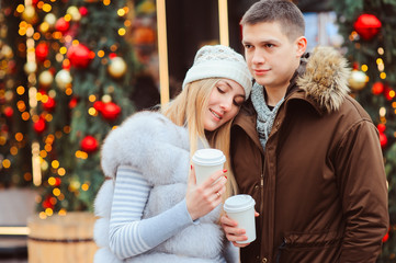 christmas portrait of happy couple with hot mulled wine or tea walking on city streets decorated for holidays, christmas trees with lights and toys on background