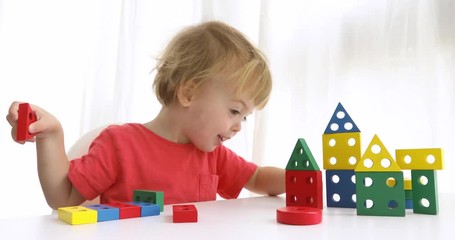 Charming infant boy sitting at table and playing with colorful constructor blocks on white background