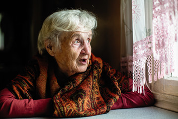 Elderly woman sitting in the house looks longingly out the window.