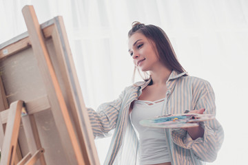 attractive artist holding palette and looking at easel in living room