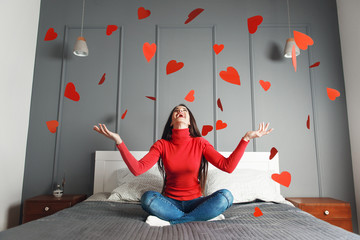 Beautiful, happy, young woman throwing red heart-shapes in the air, sitting on the bed
