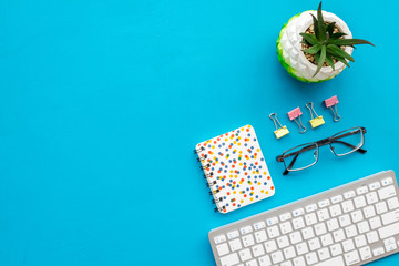 Work desk of manager. Computer keyboard, glasses, stationery, plant on blue background top view copy space