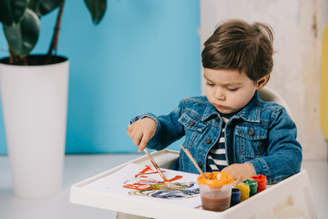 cute little boy painting with watercolor paints while sitting on highchair
