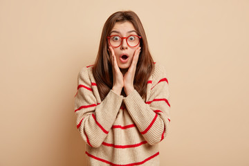 Portrait of astonished pretty young woman keeps mouth widely opened, touches cheeks, wears spectacles and sweater with red stripes, isolated over beige background. Negative emotions concept.
