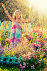 Playfull girl watering flowers with rain in the garden at summer day. Child using garden hose on sunny day at sunset lite. Little gardener playing in garden.