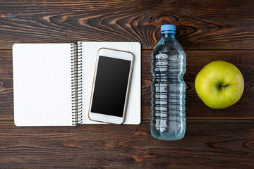 Mobile phone, apple, bottle of water and notebook on dark wooden background. 