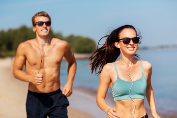 fitness, sport and technology concept - happy couple with earphones running along summer beach