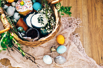 Stylish Easter eggs, easter bread cake, ham, beets, sausage, butter, green branches in wicker basket on rustic fabric with spring flowers. Happy Easter, holiday traditional feast