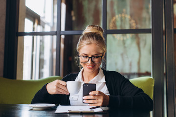 Blond business woman sitting on workspace and using laptop at office, she looks at a folder with documents, talking by phone and drinking cup of coffee.