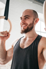 Close up view of bearded fit sportsman holding gymnastic rings at cross gym. Smiling athlete resting after training on gymnastic rings in sport hall. Vertical