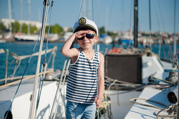 cute happy caucasian kid in captain cap and sunglasses on yacht board in summer sea port