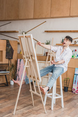excited artist sitting on high chair at easel with canvas