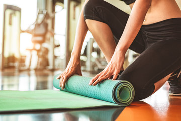 Close up of sporty woman folding yoga mattress in sport fitness gym training center background. Exercise mat rolling keeping after yoga class. Workout and sport training concept. Hands on carpet