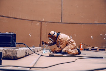 Worker in protective suit and mask crouching and welding in metal tower at construction site.