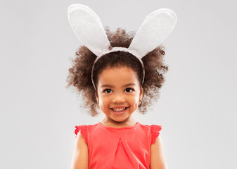 childhood, party props and easter concept - happy little african american girl wearing bunny ears headband over grey background