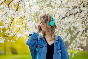 girl in a denim jacket and headphones stands near a flowering tree
