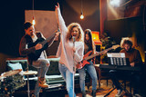 Band practice for the show. Woman with curly hair holding microphone and singing while man in background playing acoustic guitar. Home studio interior.