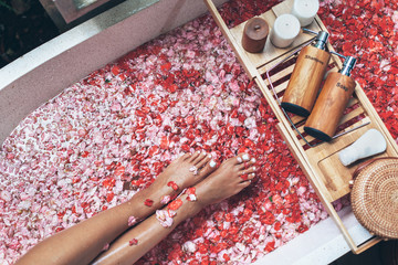 Female legs in bathtub with flower petals and beauty products on wooden tray