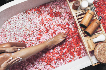 Female legs in bathtub with flower petals and beauty products on wooden tray