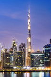 Stunning view of the illuminated Dubai skyline during sunset with the magnificent Burj Khalifa and many other buildings and skyscrapers reflected on a silky smooth water flowing in the foreground.