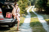 Young woman enjoying nature while sitting in the car trunk on a picturesque road in the woods