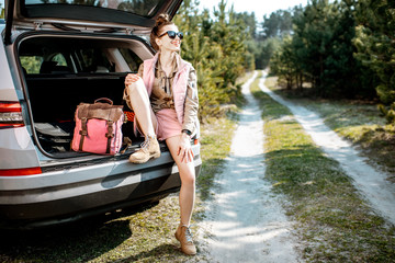 Young woman enjoying nature while sitting in the car trunk on a picturesque road in the woods