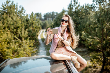Young woman enjoying a trip, photographing with phone while sitting on the car roof on a picturesque road in the woods