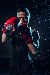 Concentrated boxer in red boxing gloves standing under water drops on black