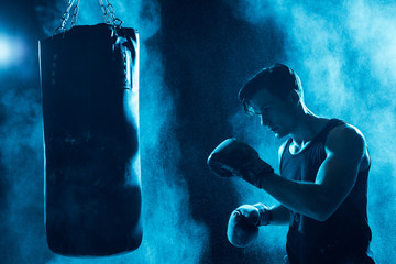 Concentrated boxer in boxing gloves training with punching bag in dark