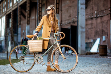 Full body portrait of a beautiful stylish woman dressed in coat standing with retro bicycle outdoors on the industrial urban background